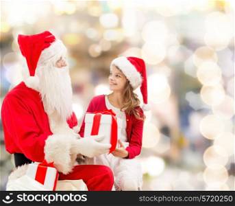 holidays, christmas, childhood and people concept - smiling little girl with santa claus and gifts over lights background