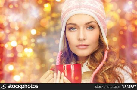 holidays, christmas, drinks and people concept - close up of young woman in winter clothes with red tea cup over lights background