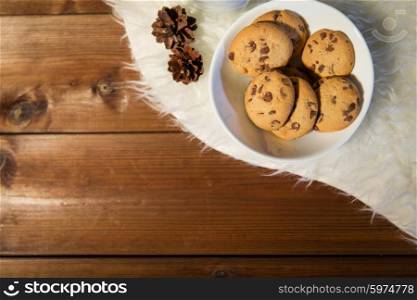 holidays, christmas, winter, advertisement and food concept - close up of cookies in bowl and cones on white fur rug on wooden board