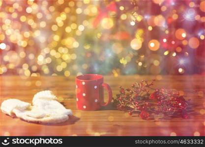 holidays, christmas, winter and drinks concept - close up of tea cup with mittens and christmas decoration on wooden table over lights