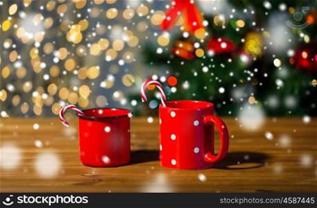 holidays, christmas, winter, food and drinks concept - close up of candy canes and cups on wooden table over lights