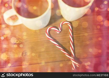 holidays, christmas, winter, food and drinks concept - close up of candy canes and cups with hot chocolate or cocoa drinks on wooden table over lights