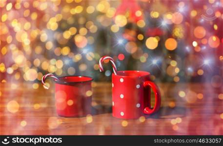 holidays, christmas, winter, food and drinks concept - close up of candy canes in cups on wooden table over lights