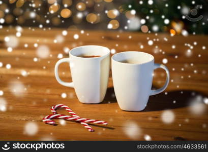 holidays, christmas, winter, food and drinks concept - close up of candy canes and cups with hot chocolate or cocoa drinks on wooden table over christmas tree lights