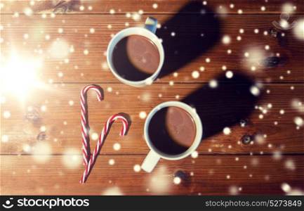holidays, christmas, winter, food and drinks concept - close up of candy canes and cups with hot chocolate or cocoa drinks on wooden table. christmas candy canes and cups on wooden table