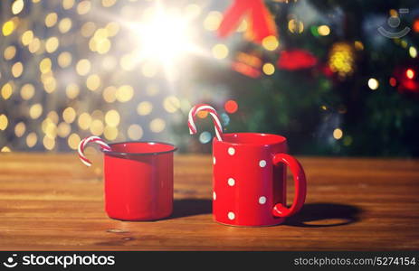 holidays, christmas, winter, food and drinks concept - close up of candy canes and cups on wooden table over lights. christmas candy canes and cups on wooden table