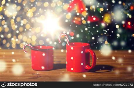 holidays, christmas, winter, food and drinks concept - close up of candy canes and cups on wooden table over lights. christmas candy canes and cups on wooden table
