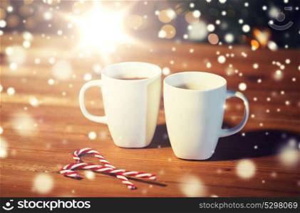 holidays, christmas, winter, food and drinks concept - close up of candy canes and cups with hot chocolate or cocoa drinks on wooden table over christmas tree lights. christmas candy canes and cups on wooden table