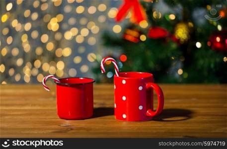 holidays, christmas, winter, food and drinks concept - close up of candy canes and cups on wooden table over lights