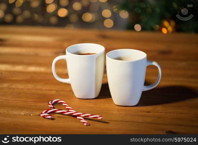 holidays, christmas, winter, food and drinks concept - close up of candy canes and cups with hot chocolate or cocoa drinks on wooden table over christmas tree lights