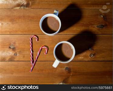holidays, christmas, winter, food and drinks concept - close up of candy canes and cups with hot chocolate or cocoa drinks on wooden table