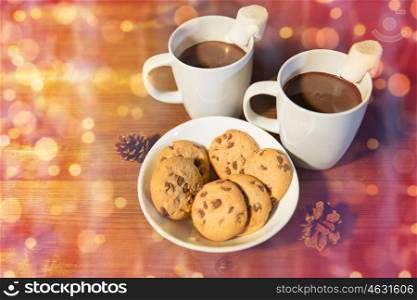 holidays, christmas, winter, food and drinks concept - close up of cups with hot chocolate or cocoa drinks and marshmallow with oat cookies on wooden table over lights