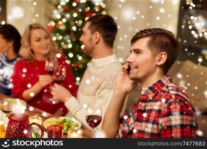holidays, communication and celebration concept - happy young man calling on smartphone and having christmas dinner with friends at home over snow. man calling on smartphone at christmas dinner