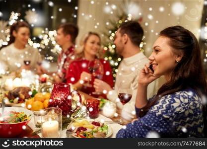holidays, communication and celebration concept - happy young woman calling on smartphone and having christmas dinner with friends at home over snow. woman calling on smartphone at christmas dinner
