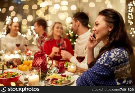 holidays, communication and celebration concept - happy young woman calling on smartphone and having christmas dinner with friends at home. woman calling on smartphone at christmas dinner