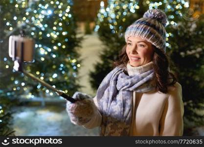 holidays, communication and people concept - portrait of beautiful happy smiling young woman with smartphone and selfie stick taking picture over christmas lights in winter city. happy woman taking christmas selfie on smartphone