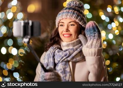 holidays, communication and people concept - portrait of beautiful happy smiling young woman with smartphone and selfie stick taking picture over christmas lights in winter city. happy woman taking christmas selfie on smartphone