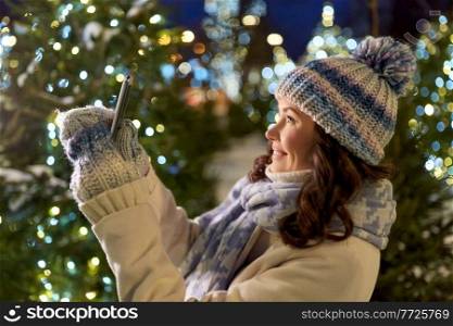 holidays, communication and people concept - portrait of beautiful happy smiling young woman with smartphone taking selfie over christmas lights in winter city. woman with smartphone taking christmas selfie
