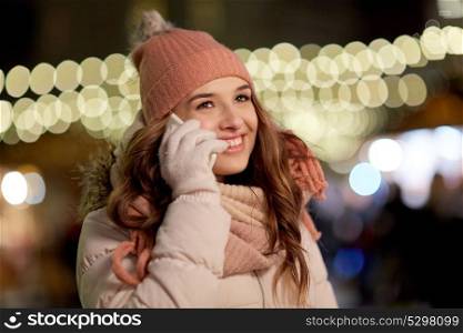 holidays, communication and people concept - portrait of beautiful happy young woman calling on smartphone over christmas lights in winter evening. happy woman calling on smartphone at christmas