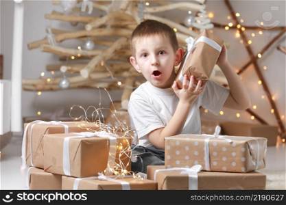 Holidays Concept. A cute little excited boy child is sitting by the wooden decorations Christmas tree and many gift boxes, holding a gift box. new Year&rsquo;s Eve and Christmas, waiting for a miracle.. Holidays Concept. A cute little excited boy child is sitting by the wooden decorations Christmas tree and many gift boxes, holding a gift box. new Year&rsquo;s Eve and Christmas, waiting for a miracle