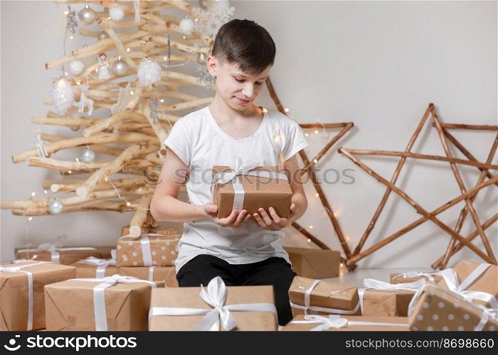Holidays Concept. A smiling excited boy child by the wooden decoration Christmas tree, in his hands he holds a big gift box. new Year’s Eve and Christmas, waiting for a miracle.. Holidays Concept. A smiling excited boy child by the wooden decoration Christmas tree, in his hands he holds a big gift box. new Year’s Eve and Christmas, waiting for a miracle