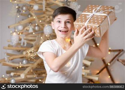 Holidays Concept. A smiling excited boyχld by the wooden decoration Christmas tree, in his hands he holds a big gift box.≠w Year’s Eve and Christmas, waiting for a mirac≤.. Holidays Concept. A smiling excited boyχld by the wooden decoration Christmas tree, in his hands he holds a big gift box.≠w Year’s Eve and Christmas, waiting for a mirac≤