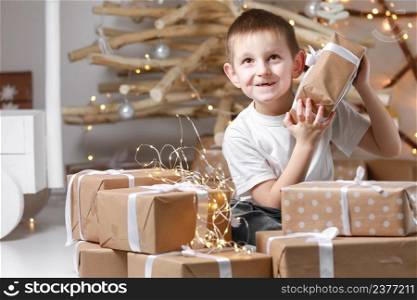 Holidays Concept. A smiling little excited boy child is sitting by the wooden decorations Christmas tree and many gift boxes, holding a gift box. new Year&rsquo;s Eve and Christmas, waiting for a miracle.. Holidays Concept. A smiling little excited boy child is sitting by the wooden decorations Christmas tree and many gift boxes, holding a gift box. new Year&rsquo;s Eve and Christmas, waiting for a miracle