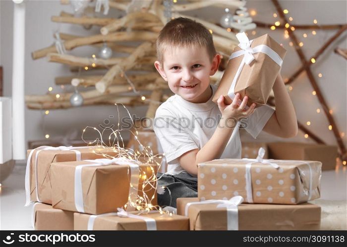 Holidays Concept. A smiling little excited boy child is sitting by the wooden decorations Christmas tree and many gift boxes, holding a gift box. new Year&rsquo;s Eve and Christmas, waiting for a miracle.. Holidays Concept. A smiling little excited boy child is sitting by the wooden decorations Christmas tree and many gift boxes, holding a gift box. new Year&rsquo;s Eve and Christmas, waiting for a miracle