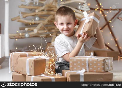 Holidays Concept. A smiling little excited boy child is sitting by the wooden decorations Christmas tree and many gift boxes, holding a gift box. new Year&rsquo;s Eve and Christmas, waiting for a miracle.. Holidays Concept. A smiling little excited boy child is sitting by the wooden decorations Christmas tree and many gift boxes, holding a gift box. new Year&rsquo;s Eve and Christmas, waiting for a miracle