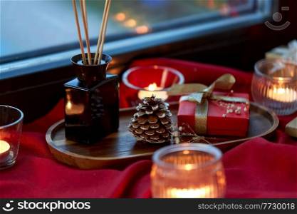 holidays, decoration and celebration concept - christmas gift, candles and aroma reed diffuser on red tablecloth on window sill at home. christmas gift, candles, reed diffuser on window
