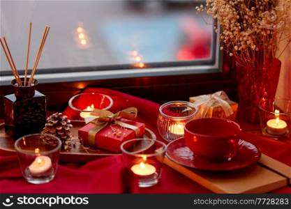 holidays, decoration and celebration concept - christmas gift, coffee cup cup, candles and aroma reed diffuser on red tablecloth on window sill at home. christmas gift, candles and cup on window sill