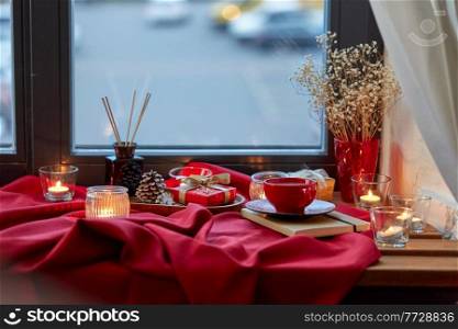 holidays, decoration and celebration concept - christmas gift, cup of coffee, candles and aroma reed diffuser on red tablecloth on window sill at home. christmas gift, candles and coffee on window sill