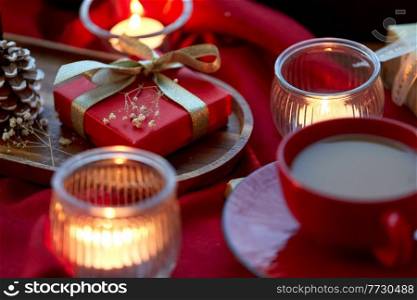 holidays, decoration and celebration concept - close up of christmas gift, cup of coffee and candles on red tablecloth on window sill at home. christmas gift, candles and coffee on window sill