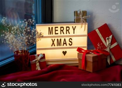 holidays, decoration and celebration concept - close up of merry christmas greeting on light box and gift boxes on red tablecloth on window sill at home. merry christmas on light box and gifts on window