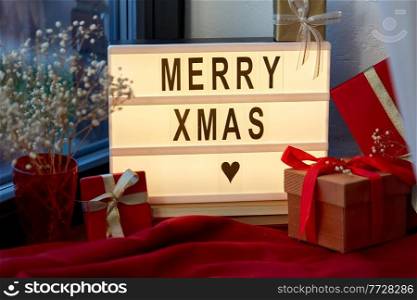 holidays, decoration and celebration concept - merry christmas greeting on light box and gift boxes on red tablecloth on window sill at home. merry christmas on light box and gifts on window