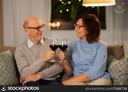 holidays, drinks and people concept - happy smiling senior couple toasting glasses of red wine at home in evening. happy senior couple with glasses of red wine