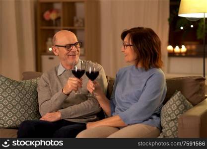 holidays, drinks and people concept - happy smiling senior couple toasting glasses of red wine at home in evening. happy senior couple with glasses of red wine