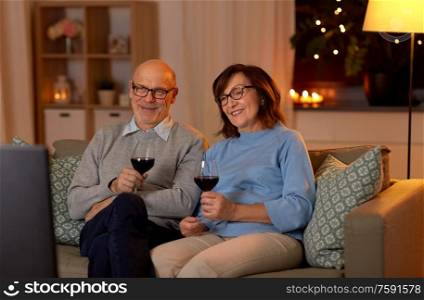 holidays, drinks and people concept - happy smiling senior couple with glasses of red wine watching tv at home in evening. happy senior couple drink red wine and watch tv