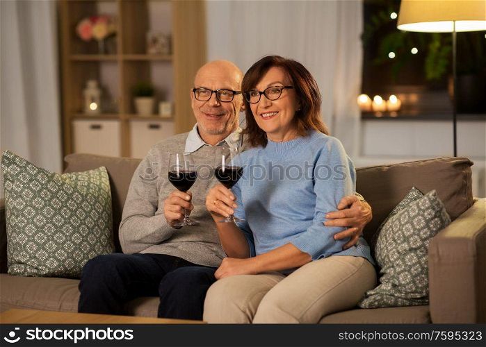 holidays, drinks and people concept - happy smiling senior couple with glasses of red wine at home in evening. happy senior couple with glasses of red wine