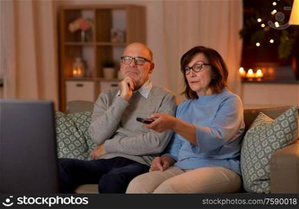holidays, drinks and people concept - senior couple with remote control watching tv at home in evening. senior couple watching tv at home in evening