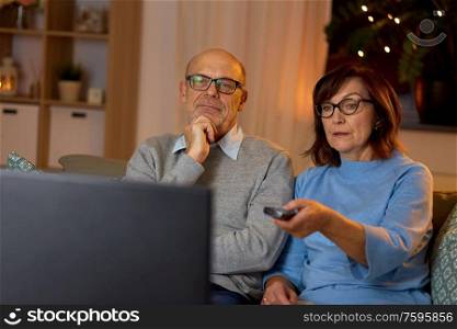holidays, drinks and people concept - senior couple with remote control watching tv at home in evening. senior couple watching tv at home in evening