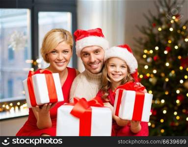 holidays, family and celebration concept - happy mother, father and daughter with gifts at home over christmas tree lights background. happy family with christmas gifts at home