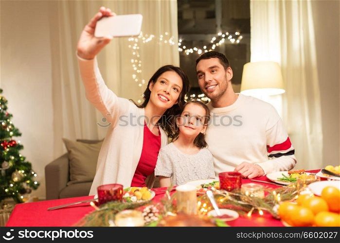 holidays, family and celebration concept - happy mother, father and little daughter having christmas dinner and taking selfie picture by smartphone at home. happy family taking selfie at christmas dinner