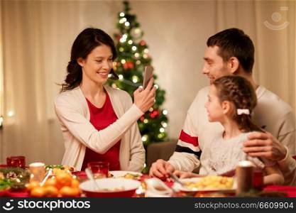 holidays, family and celebration concept - happy mother, father and little daughter having christmas dinner and taking picture by smartphone at home. happy family taking picture at christmas dinner