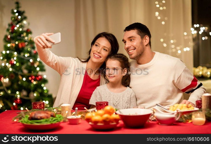 holidays, family and celebration concept - happy mother, father and little daughter having christmas dinner and taking selfie picture by smartphone at home. happy family taking selfie at christmas dinner