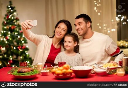 holidays, family and celebration concept - happy mother, father and little daughter having christmas dinner and taking selfie picture by smartphone at home. happy family taking selfie at christmas dinner