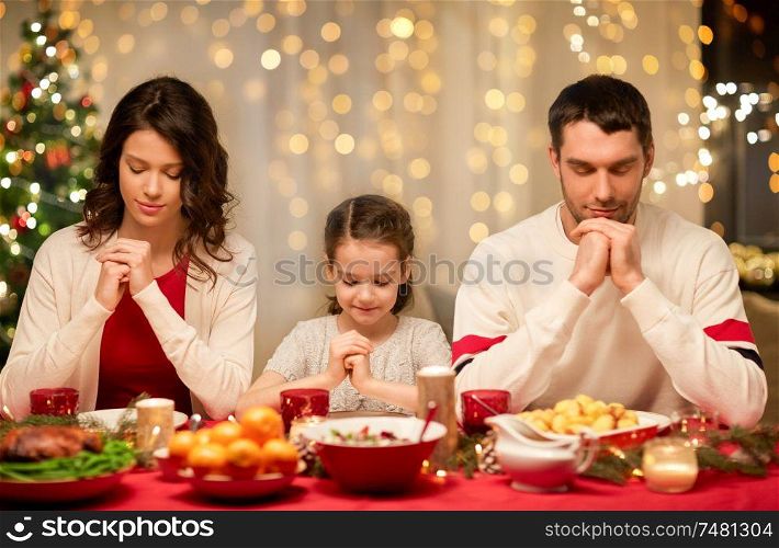 holidays, family and celebration concept - happy mother, father and little daughter having christmas dinner and praying before meal at home. family praying before meal at christmas dinner