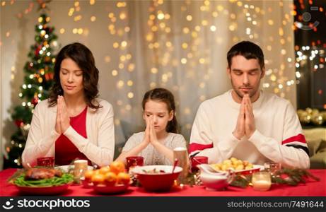 holidays, family and celebration concept - happy mother, father and little daughter having christmas dinner and praying before meal at home. family praying before meal at christmas dinner