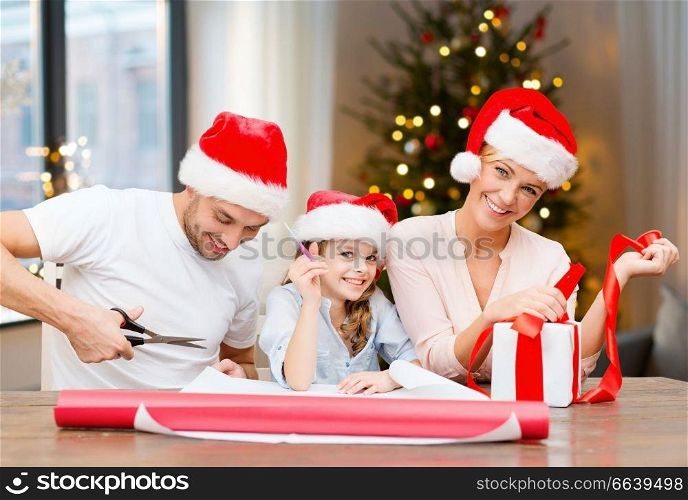 holidays, family and craft concept - happy mother, father and daughter wrapping gifts at home over christmas tree background. happy family wrapping christmas gifts at home