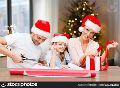 holidays, family and craft concept - happy mother, father and daughter wrapping gifts at home over christmas tree background. happy family wrapping christmas gifts at home
